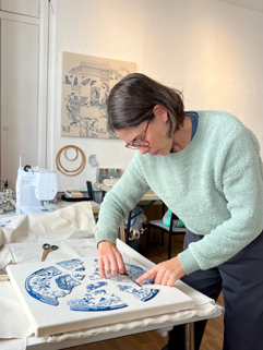 Image description for those with visual impairments: Artist Deniz Kurdak in her studio cutting the loose threads of an embroidered artwork with fragments of a blue and white chinoiserie-style design on canvas. The design resembles broken pieces of pottery, reconstructed in a circular form. In the background, there are a sewing machine, some tools and fabrics, embroidery hoops, and a finished artwork with a similar pottery pattern hanging on the wall.