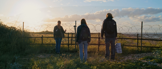 A family of three people stand with their backs to the camera looking through a razor wire to a border fence. The sun is setting.